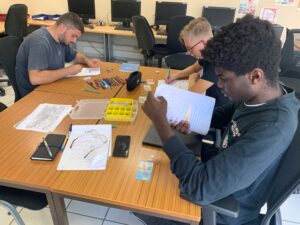 Trois jeunes hommes sont assis autour d'une table en bois clair, concentrés sur des cahiers et feuilles de travail. La table est couverte de crayons de couleur, pièces de monnaie, un téléphone et des notes. En arrière-plan, plusieurs ordinateurs éteints et des chaises noires complètent la scène d’un espace d’étude.
