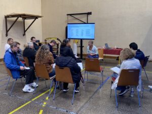 Une femme souriante devant un public de 10 personnes. Derrière elle, on voit un écran avec un diaporama - conférence lors du forum de l'emploi et de l'inclusion du COS CRPF.