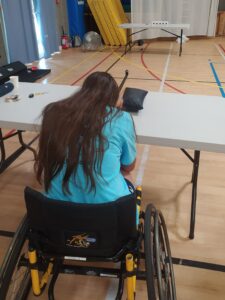 Une participante des Olympiades de LADAPT en fauteuil roulant, vue de dos, s'exerce au tir de précision avec une carabine posée sur une table dans un gymnase.